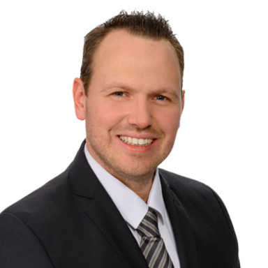 Mann in Anzug mit Krawatte, lächelnd vor weißem Hintergrund.