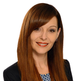 Frau mit langen, braunen Haaren und einem blauen Musterblazer, lächelt freundlich.