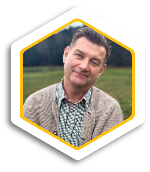 Mann mit kurzem Haar, beige Strickjacke und grünem Hemd, steht vor einer Wiese.