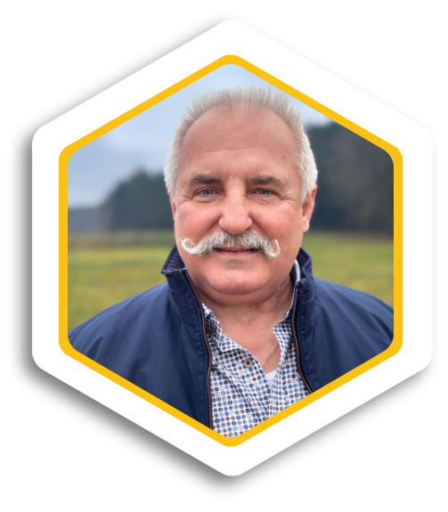 Älterer Mann mit Schnurrbart, lächelt vor einer grünen Landschaft.