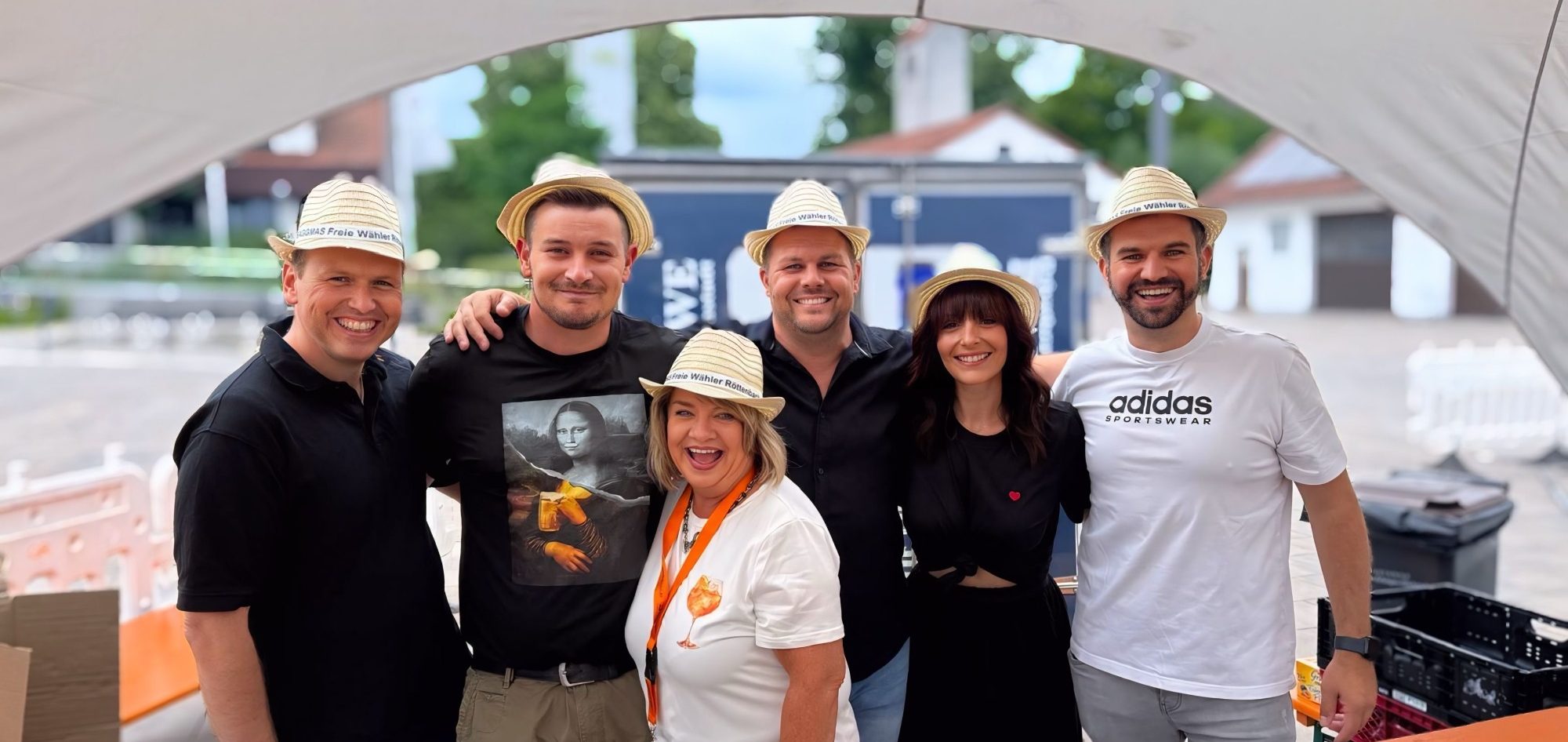 Sechs Personen in Hüten, lächeln und stehen unter einem Zeltdach.