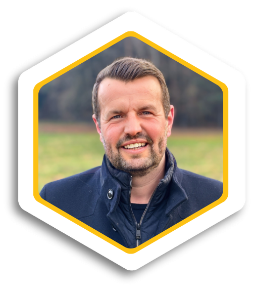 Mann mit Bart und lächelndem Gesicht, trägt eine dunkelblaue Jacke, Hintergrund mit Wiese.