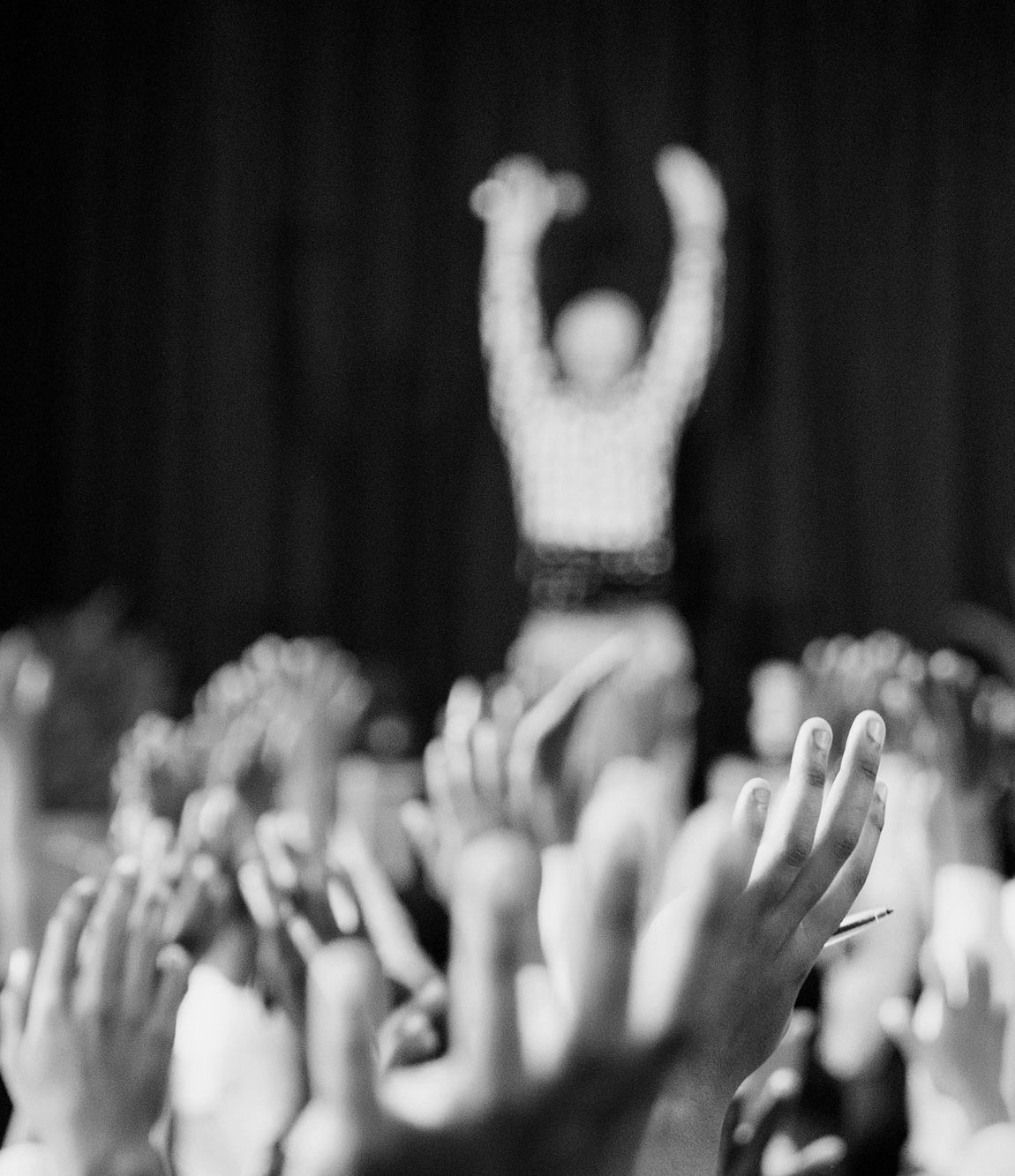Menschenmenge hebt die Hände, während eine Person im Hintergrund auf der Bühne steht.