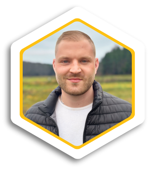 Junger Mann mit kurzen Haaren, lächelt vor einer landschaftlichen Kulisse.