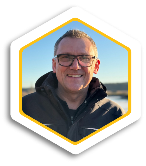 Lächelnder Mann mit Brille, in Outdoor-Bekleidung vor einem Landschaftshintergrund.