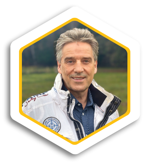 Mann mit grauen Haaren, lächelnd, in einem weißen Jackett vor einer grünen Landschaft.