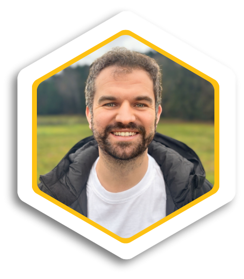 Lächelnder Mann mit Bart, trägt ein weißes T-Shirt, vor einer grünen Wiese.